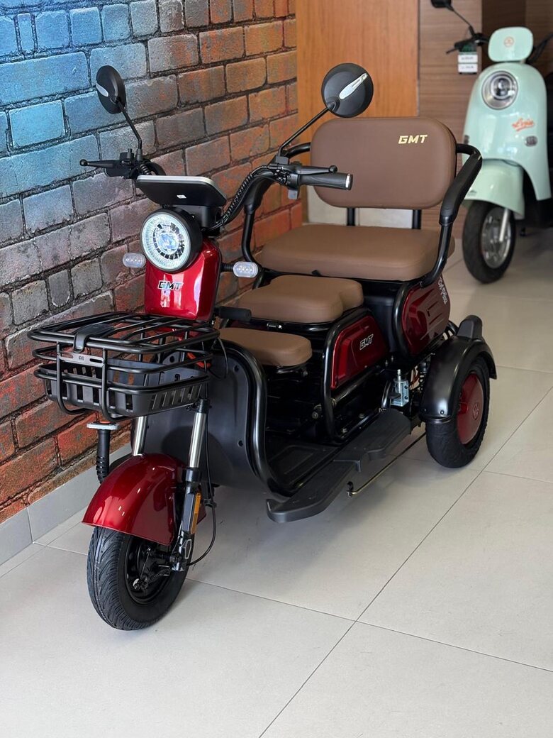 Triciclo elétrico vermelho com dois bancos e cesta, dentro da loja da KM